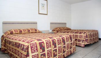 The image shows a simple room with two beds, each covered with a red and gold patterned bedspread, and a framed picture on the wall.