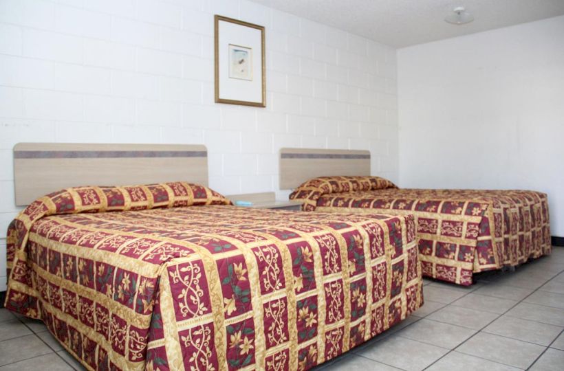 The image shows a simple room with two beds, each covered with a red and gold patterned bedspread, and a framed picture on the wall.