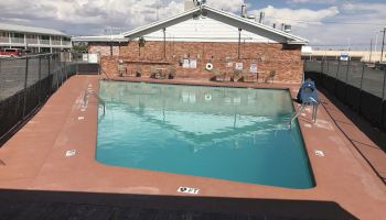 This image shows an outdoor swimming pool with a fence around it, a brick building in the background, and a clear sky with clouds.