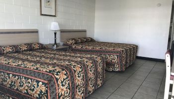 A hotel room with two double beds, patterned bedspreads, a nightstand with a lamp, a picture on the wall, tile flooring, and chairs on the right.