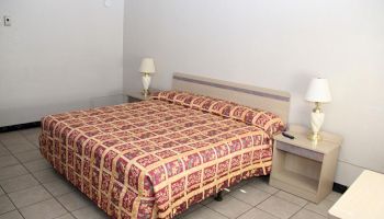 The image shows a simple hotel room with a double bed covered in a patterned bedspread, two bedside tables each with a lamp, and tiled flooring.