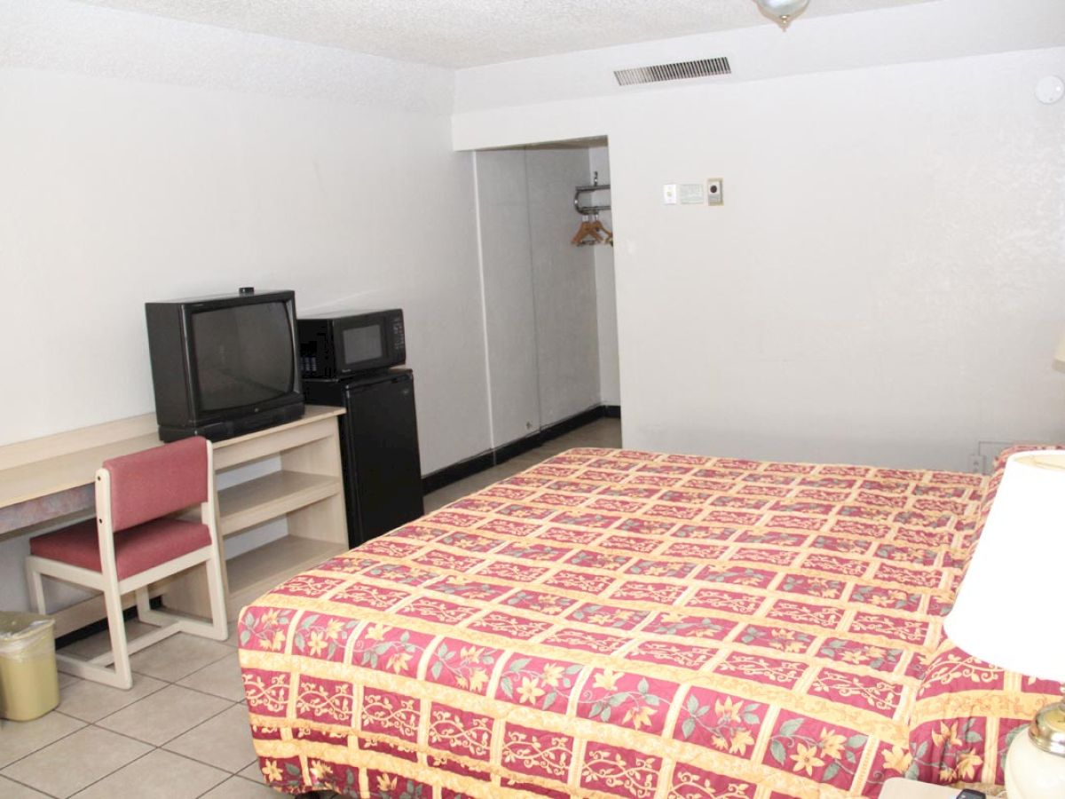 The image shows a motel room with a bed, desk, chair, TV, mini-fridge, microwave, and a closet space with hangers, featuring simple furnishings.