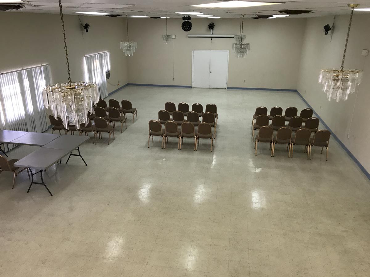 A small hall with rows of chairs arranged facing a wall with closed doors, alongside folded tables and elegant chandeliers hanging from the ceiling.