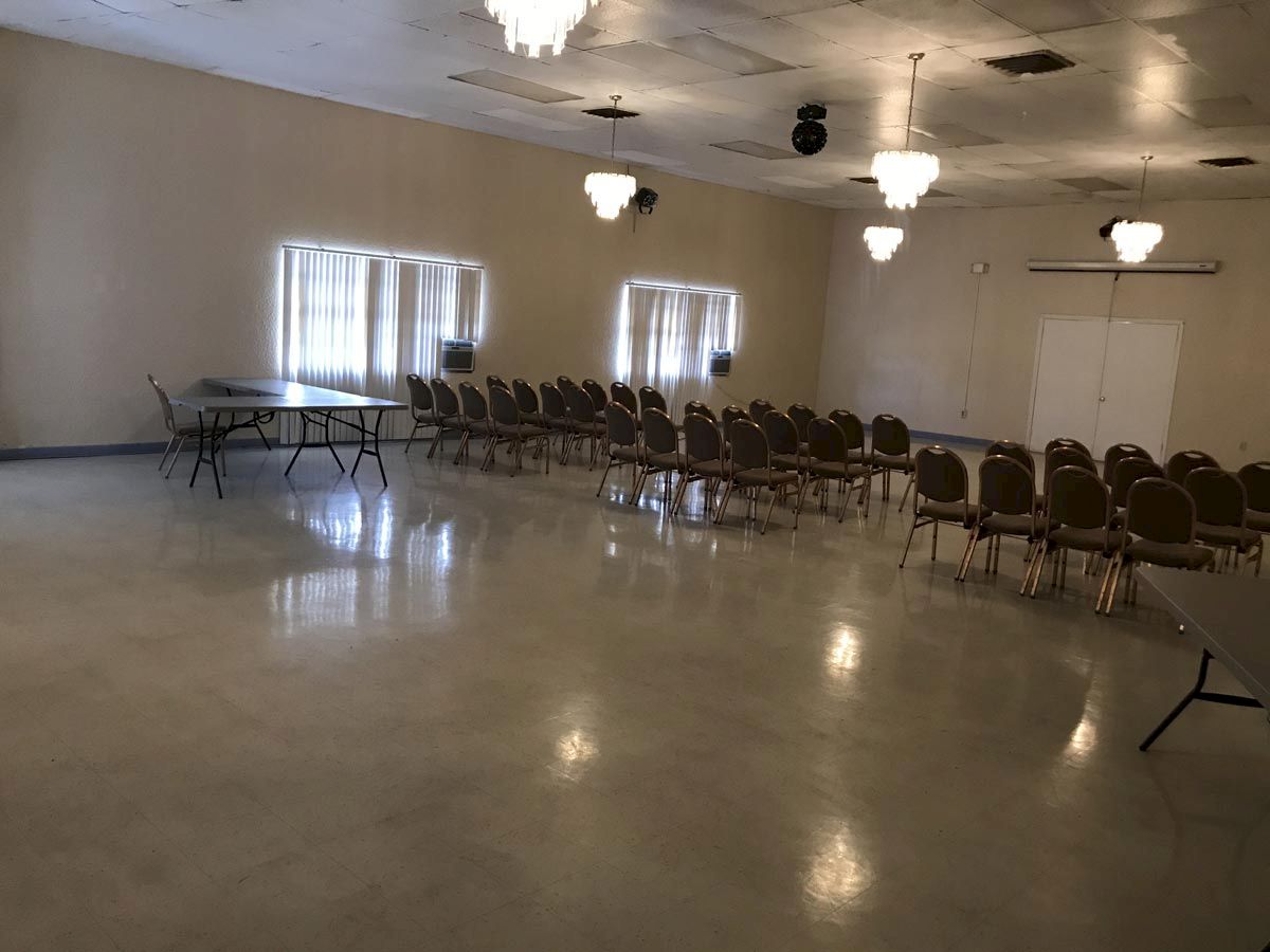 The image shows an empty room with rows of chairs arranged on one side and a few folding tables against the wall, illuminated by chandeliers.