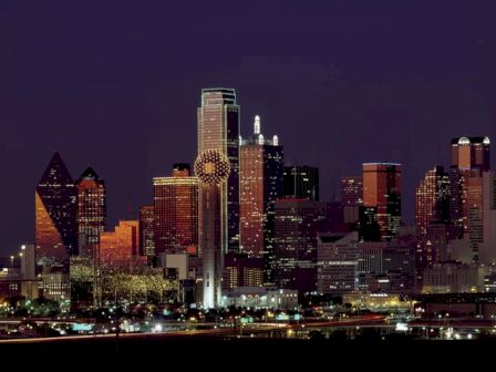 A city skyline at night, with illuminated buildings and a distinctive lit structure resembling a ball atop a tower in the foreground.