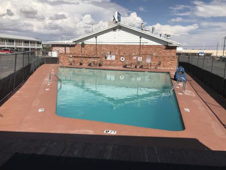 The image shows an outdoor swimming pool surrounded by a fence, with a brick building in the background and minimalistic poolside amenities.