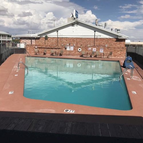 An empty outdoor swimming pool with clear water, surrounded by a brick wall and buildings in the background. The area is enclosed by a fence.