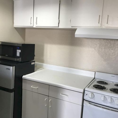 A small kitchen with white cabinets, a compact fridge, a microwave on a stand, a countertop, and a white stove with a range hood ending the sentence.