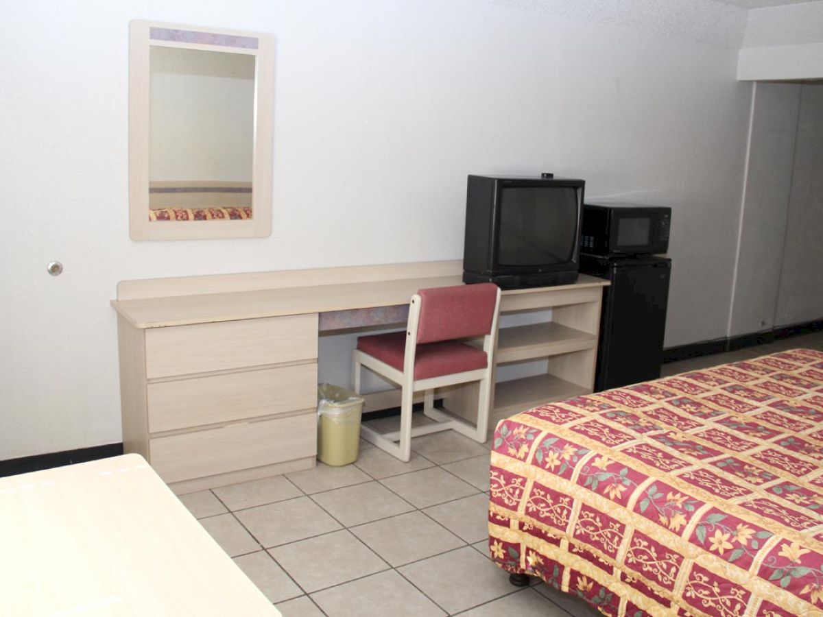 The image shows a hotel room with a bed, a desk with a mirror, a chair, a TV, a microwave, and a refrigerator, with tile flooring ending the sentence.