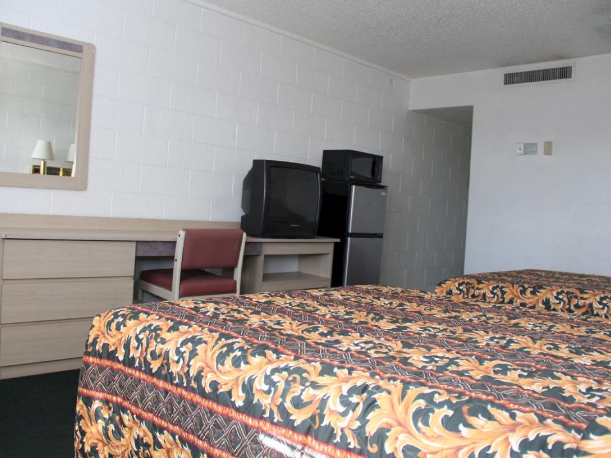 A hotel room with two beds, a small table with a chair and mirror, a TV, and a mini-fridge is seen in the image.