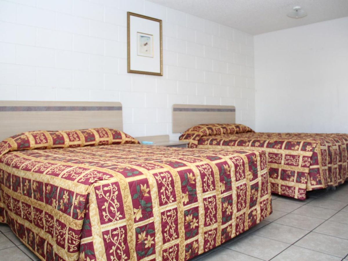 The image shows a hotel room with two double beds covered in patterned bedspreads, a simple frame on the wall, and tiled flooring.