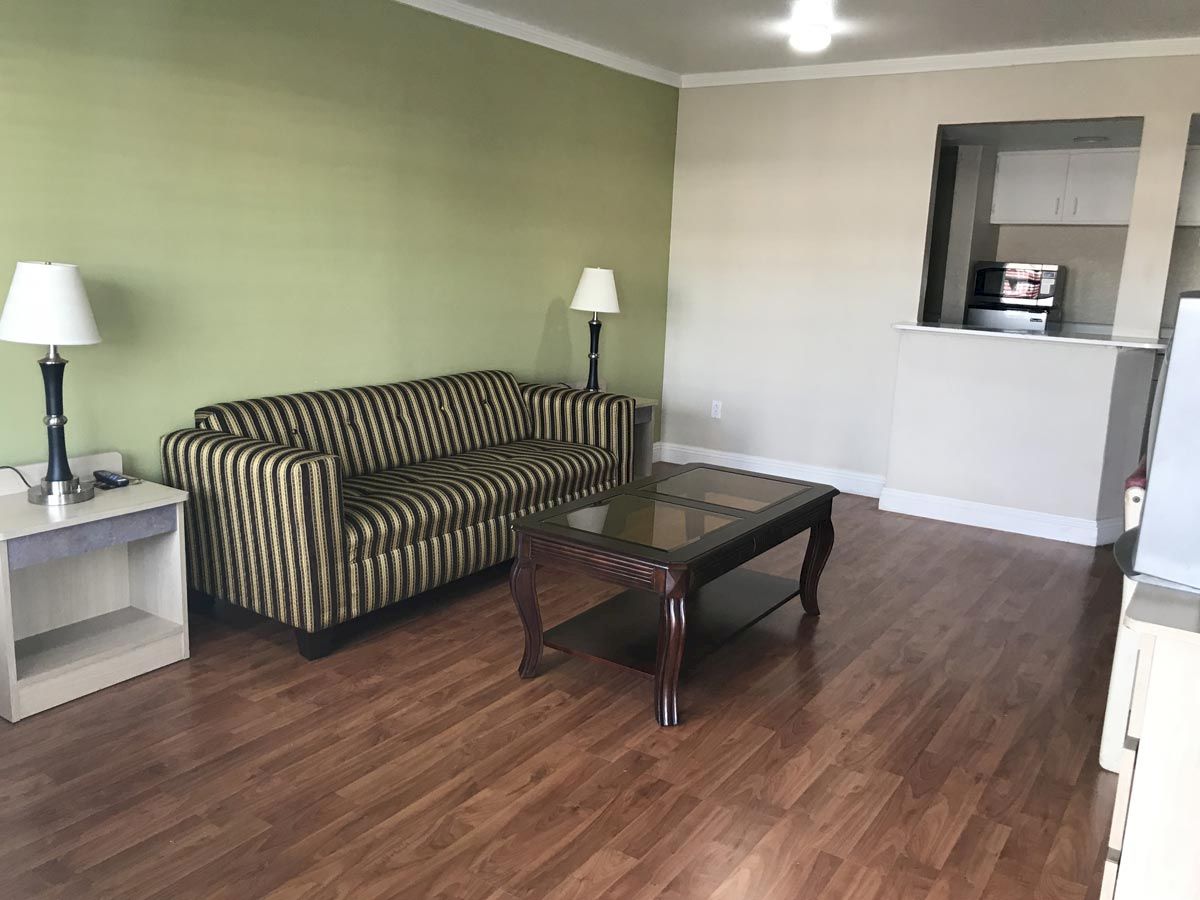 The image shows a living room with a striped sofa, two side tables with lamps, a glass coffee table, and wooden flooring, leading to a kitchen area.