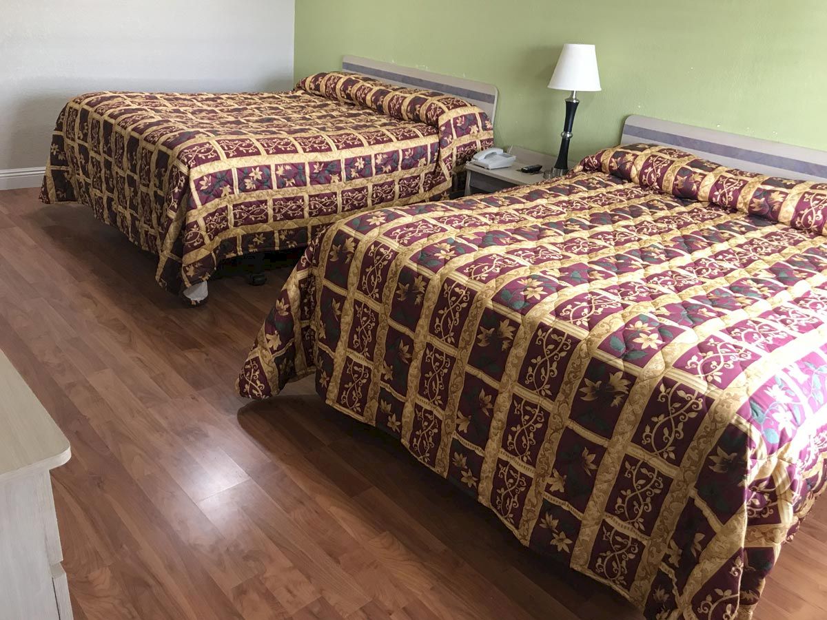 The image shows a hotel room with two double beds, both with patterned bedspreads, a nightstand with a lamp between them, and a wooden floor.