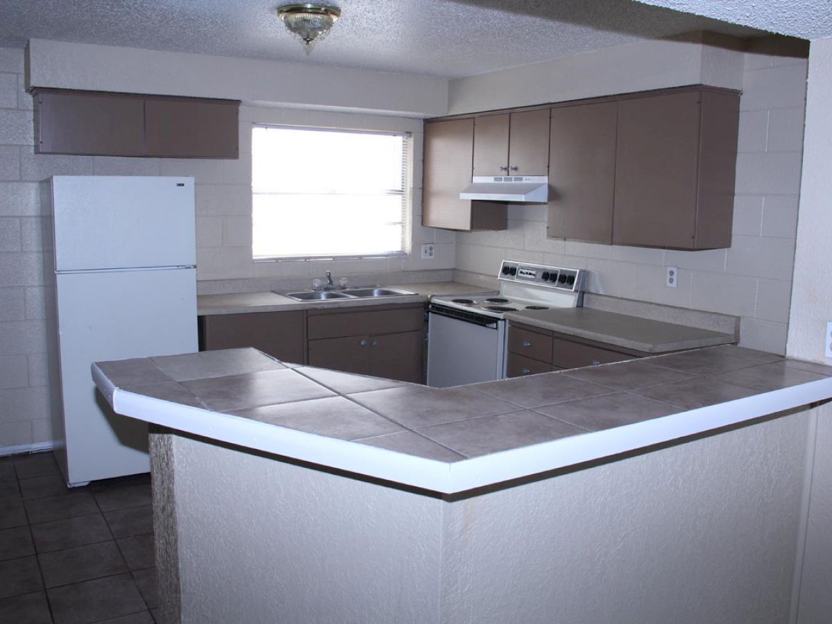 A simple kitchen with a white refrigerator, stove, dishwasher, sink, and brown cabinets. A tile countertop forms a wrap-around bar.