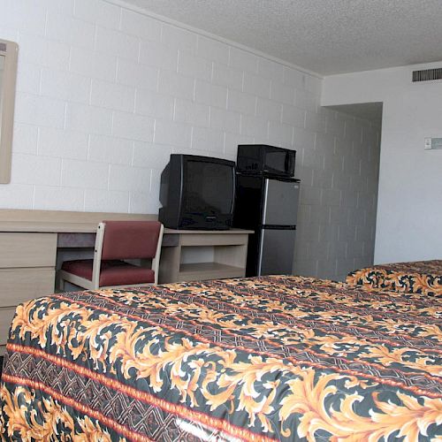 A hotel room with two beds, a desk with a mirror, a chair, a TV, and a mini-fridge. The beds have patterned bedspreads.