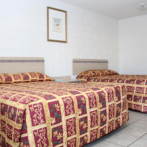 The image shows a room with two beds featuring matching patterned bedspreads, a picture on the wall, and tiled flooring.