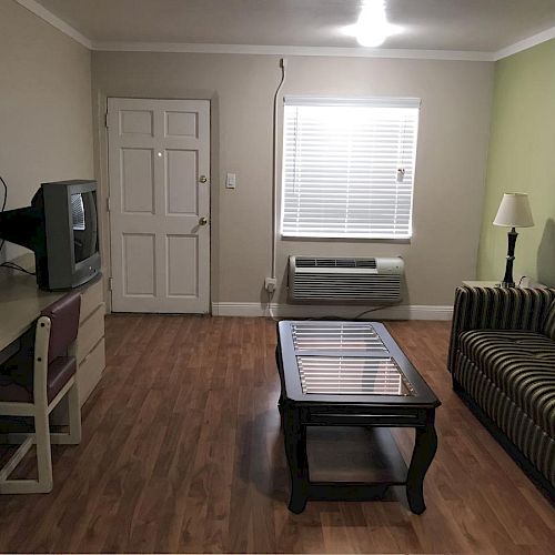 A modest living room with a TV on a desk, striped sofa, coffee table, lamp, window, door, and an air conditioning unit on a wooden floor.