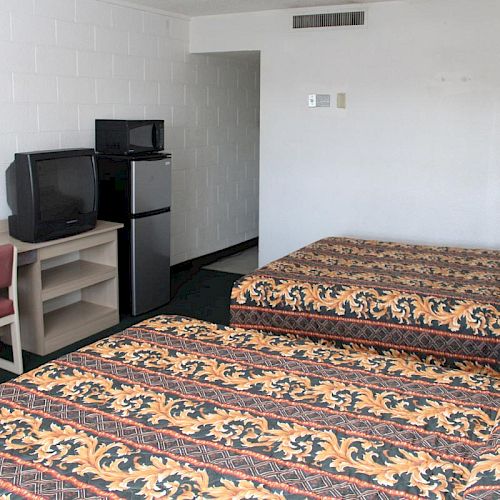 A hotel room with two double beds, a desk with a chair, a TV atop a shelf, and a mini-fridge. The beds have patterned bedspreads.