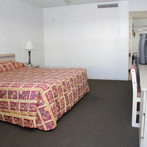 A simple hotel room with a bed, TV, lamp, and desk. There is a window on the left side, and a closet in the background.