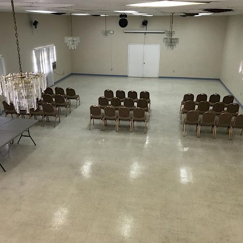 An empty room with rows of chairs set up facing the front, a few tables on the left side, and chandeliers hanging from the ceiling.