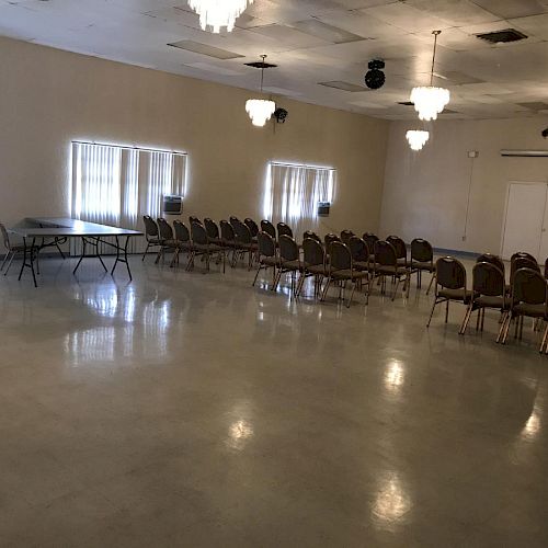 This image shows an empty room with rows of chairs and a few tables set up, likely for a meeting or event. The room has chandeliers and windows.