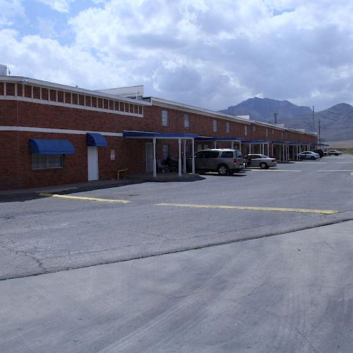 The image shows a brick building with blue awnings, some parked vehicles, a clear sky with clouds, and mountains in the background, ending the sentence.