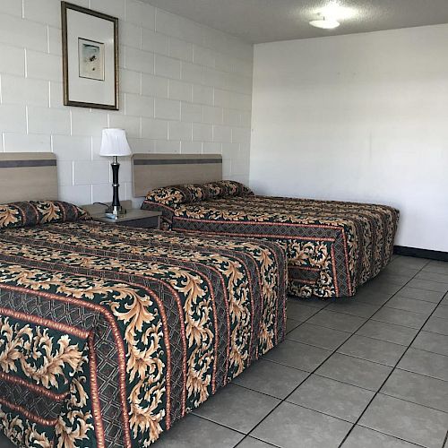 The image depicts a hotel room with two double beds, a nightstand, a lamp, and a framed picture on the wall. The floor is tiled.