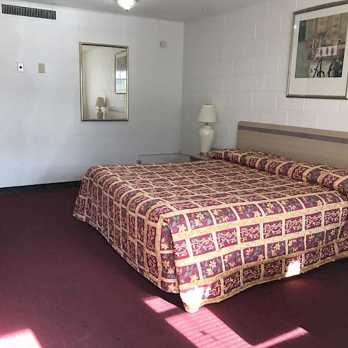 A hotel room with a large bed, a bedside table with a lamp, a framed picture on the wall, a mirror, and a chair by a desk; carpeted floor.