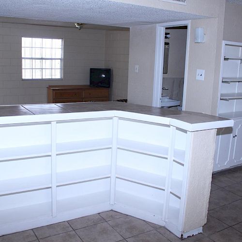 The image shows a sparsely furnished room with white shelves, a kitchen counter, tiled flooring, and a window in the background.