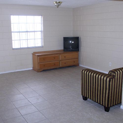 A sparsely furnished room with a small TV on a wooden dresser, a striped armchair, and a tiled floor, near a window with natural light ending the sentence.