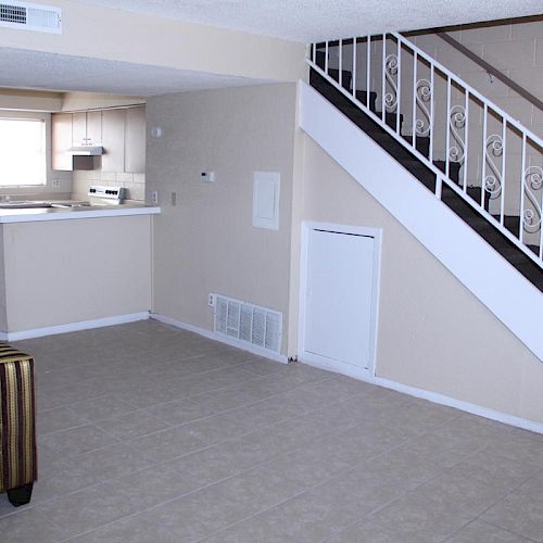 A living room with a striped armchair, staircase, and an open kitchen in the background, featuring light-colored walls and tiled flooring.