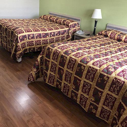 The image shows a hotel room with two double beds, colorful bedspreads, a nightstand in between, a lamp, and hardwood floors.