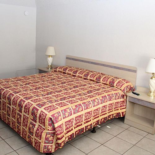 The image shows a small hotel room with a king-size bed, covered in a patterned bedspread, two bedside tables with lamps, and a tiled floor.