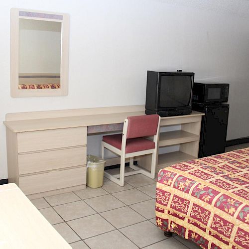 A simple hotel room with a bed, desk, chair, TV, mini-fridge, microwave, mirror, and trash bin, all arranged neatly.