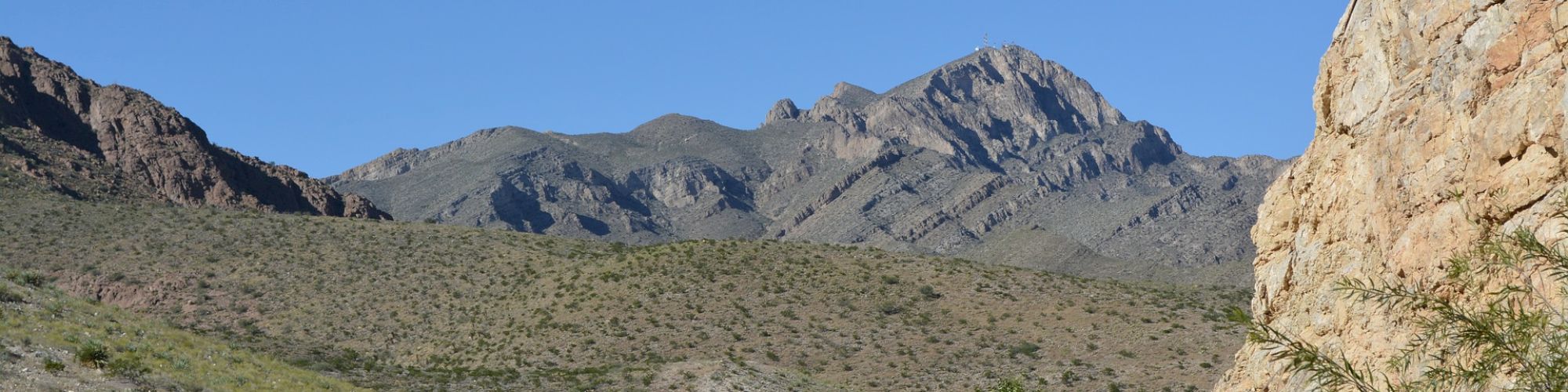 A road winds through a rocky desert landscape with sparse vegetation and a mountain range in the background under a clear blue sky.