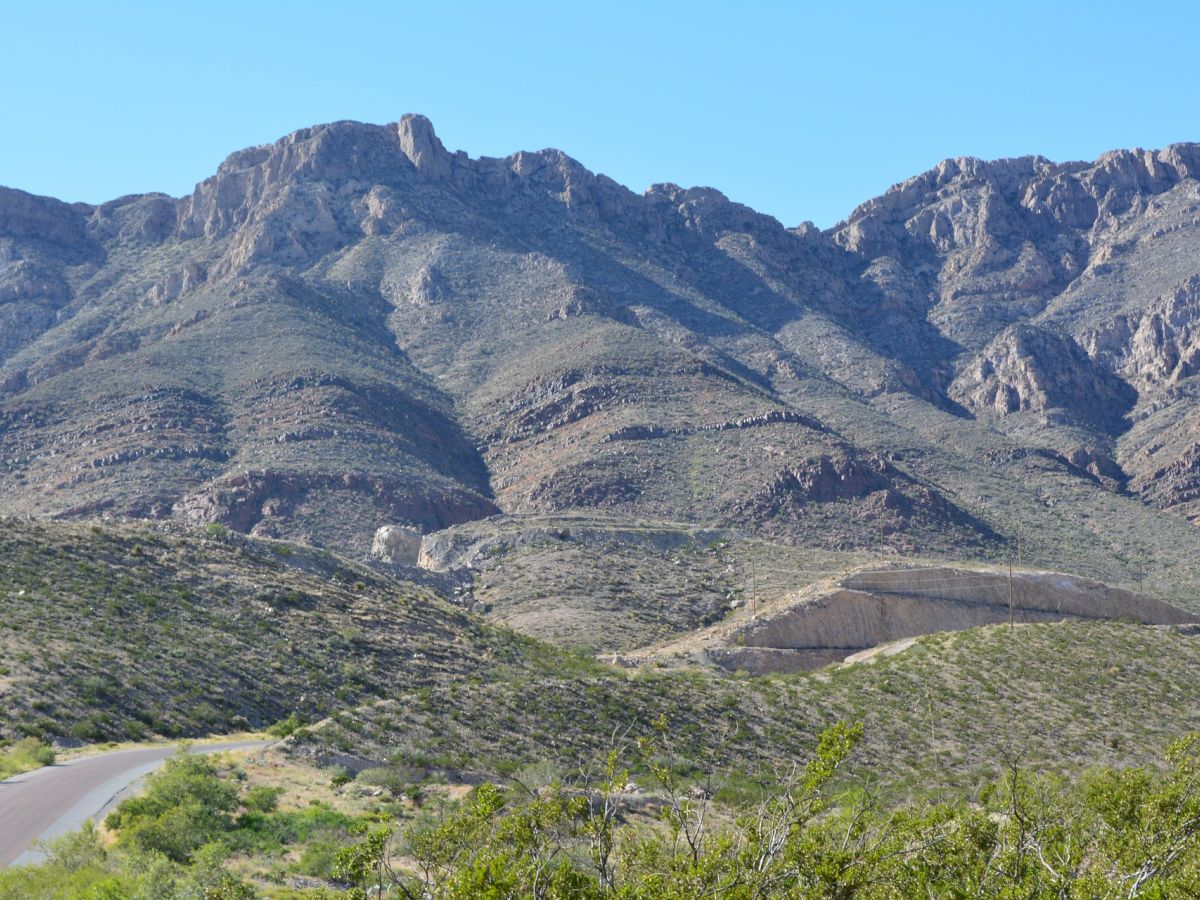 This image shows a mountainous landscape with rocky, rugged terrain, a clear blue sky, and a road winding through the foreground.