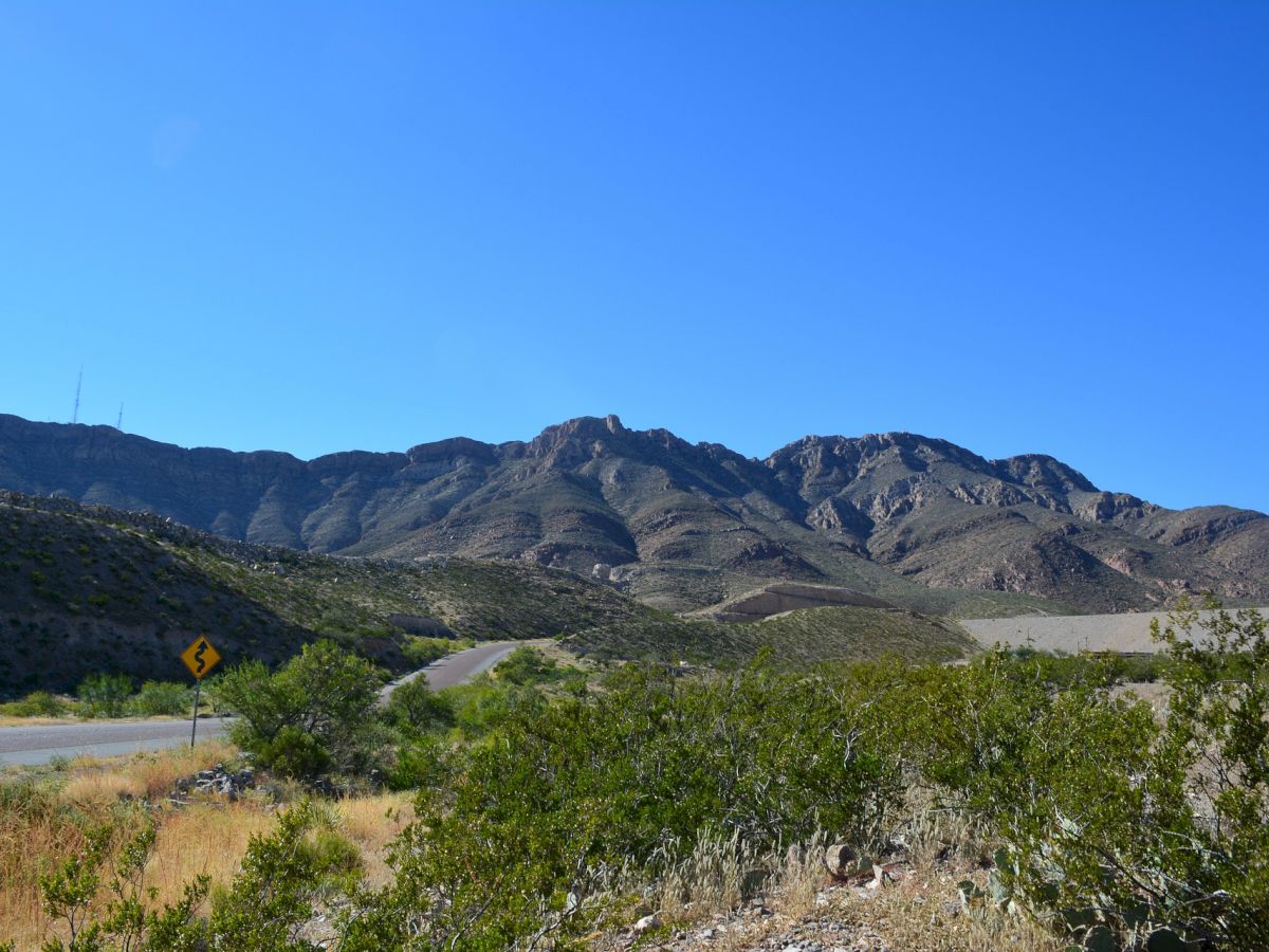 A scenic landscape with a clear blue sky, mountains, and greenery by the roadside. A caution road sign is visible on the left side of the image.