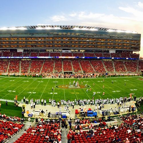 A large football stadium filled with fans, teams on the field, and bright lights as a game is underway during what appears to be early evening.