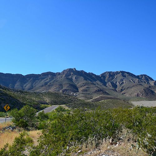 A scenic landscape features a mountain range with a clear blue sky, a winding road, and sparse vegetation in the foreground.