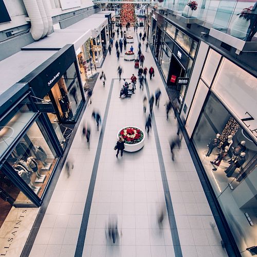 A busy indoor shopping mall with people walking past stores like Zara and All Saints. The image is taken from above, showing a central walkway.